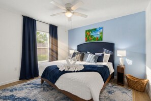 Une chambre à coucher avec un lit, un ventilateur de plafond, une fenêtre avec des rideaux, un tableau au mur et une lampe de chevet.