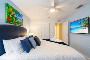 Ein Hotelzimmer mit Bett, Deckenventilator, einem Vogelgemälde und einem Fernseher, der eine Strandansicht zeigt.