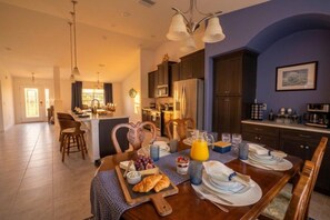 Uma sala de jantar com mesa de madeira posta para o café da manhã, cozinha ao fundo e vista para a sala de estar.