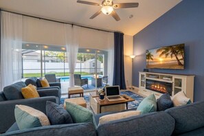 A modern living room with a sofa set, a fireplace, and a large window overlooking a pool. - Heated Pool | Massage Chair| 3 Kings| 16 Min Beach (Palm Coast)