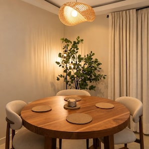 A round wooden dining table with four chairs, a woven pendant light, and a potted plant. - Selah Oasis - Kingston 2BR, 3 Beds, Sleeps 5 ~ Pool, Parking & Central Location (Kingston)