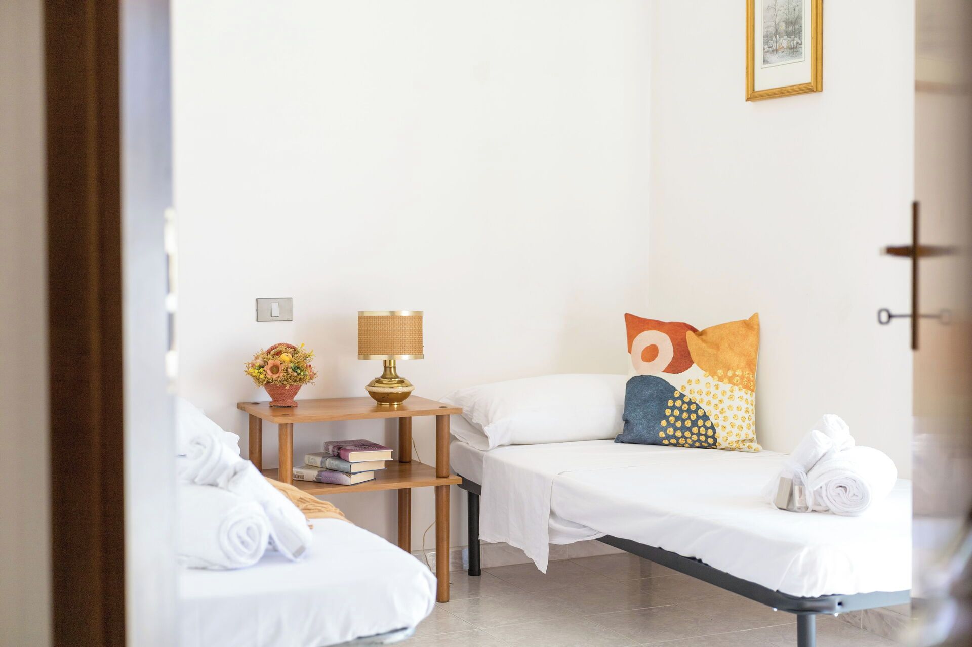 A hotel room with two single beds, a bedside table with a lamp, a small shelf with books, and a wall-mounted picture.