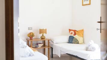 A hotel room with two single beds, a bedside table with a lamp, a small shelf with books, and a wall-mounted picture.