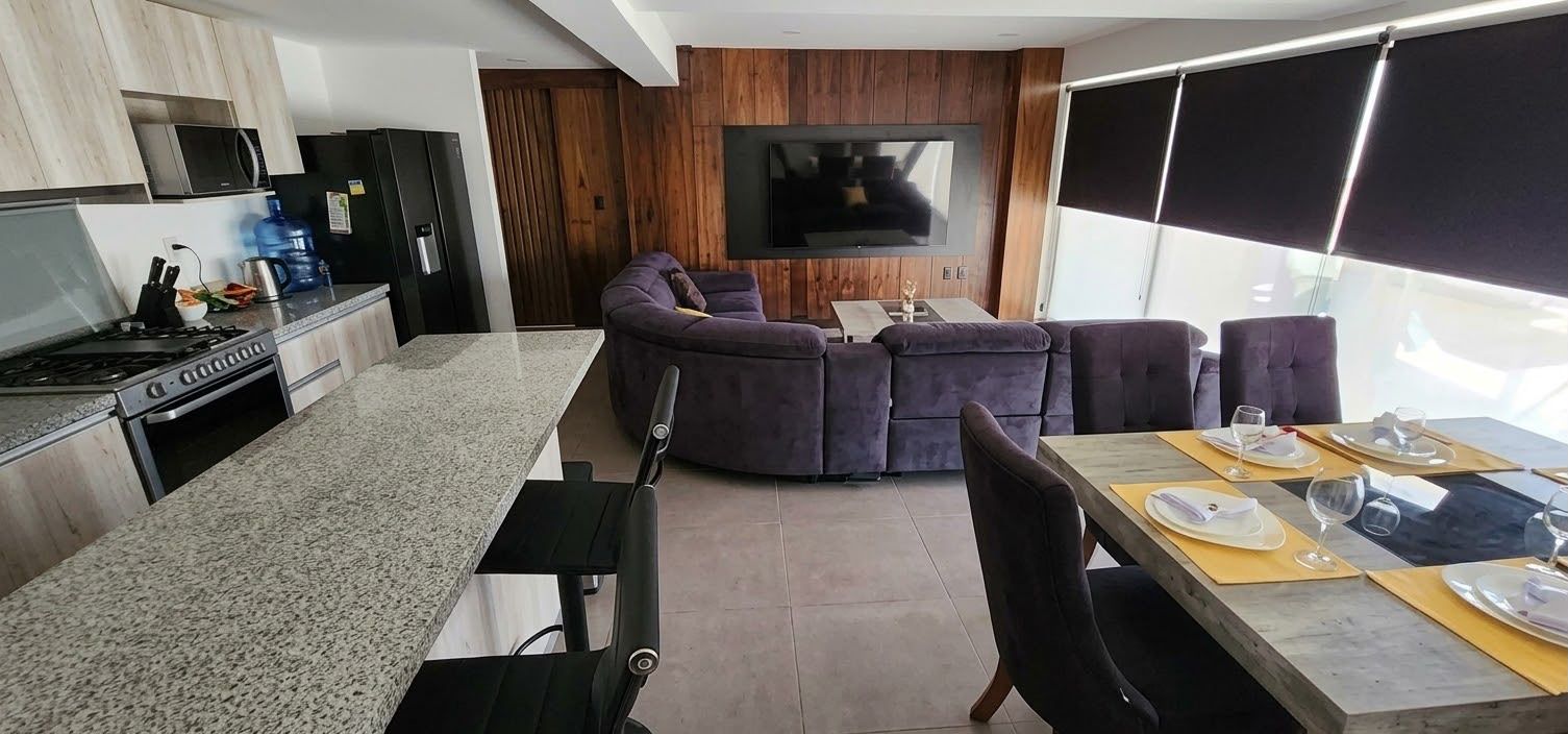 Eine moderne Küche mit Gasherd, Mikrowelle und einem großen Wasserspender. Es gibt eine Essecke mit Tisch für vier Personen, eine Couch und einen flachbildfernseher an der Wand.