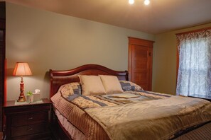 Una cama bien hecha con cabecera de madera, una lámpara de noche, una mesita de noche con caja de pañuelos y una ventana con cortinas florales.