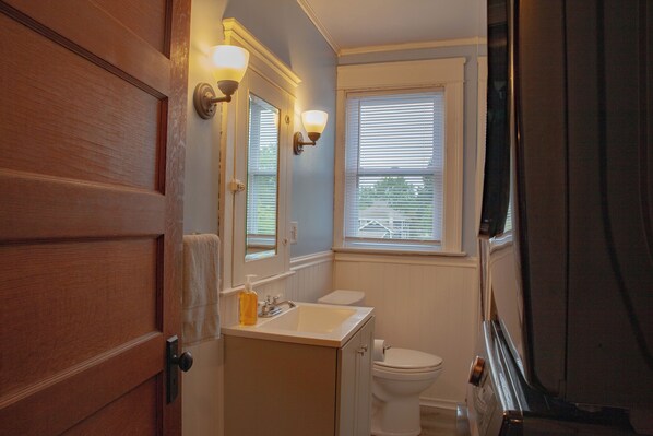 A bathroom with a wooden door, a white sink, a mirror with wall sconces, a toilet, and a window with blinds.