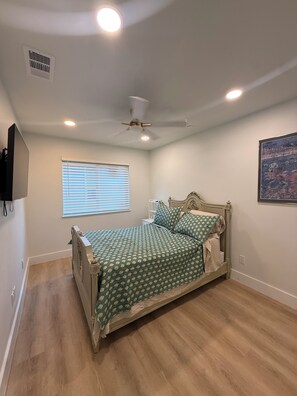 A bedroom with a bed, ceiling fan, window with blinds, and a mounted TV.