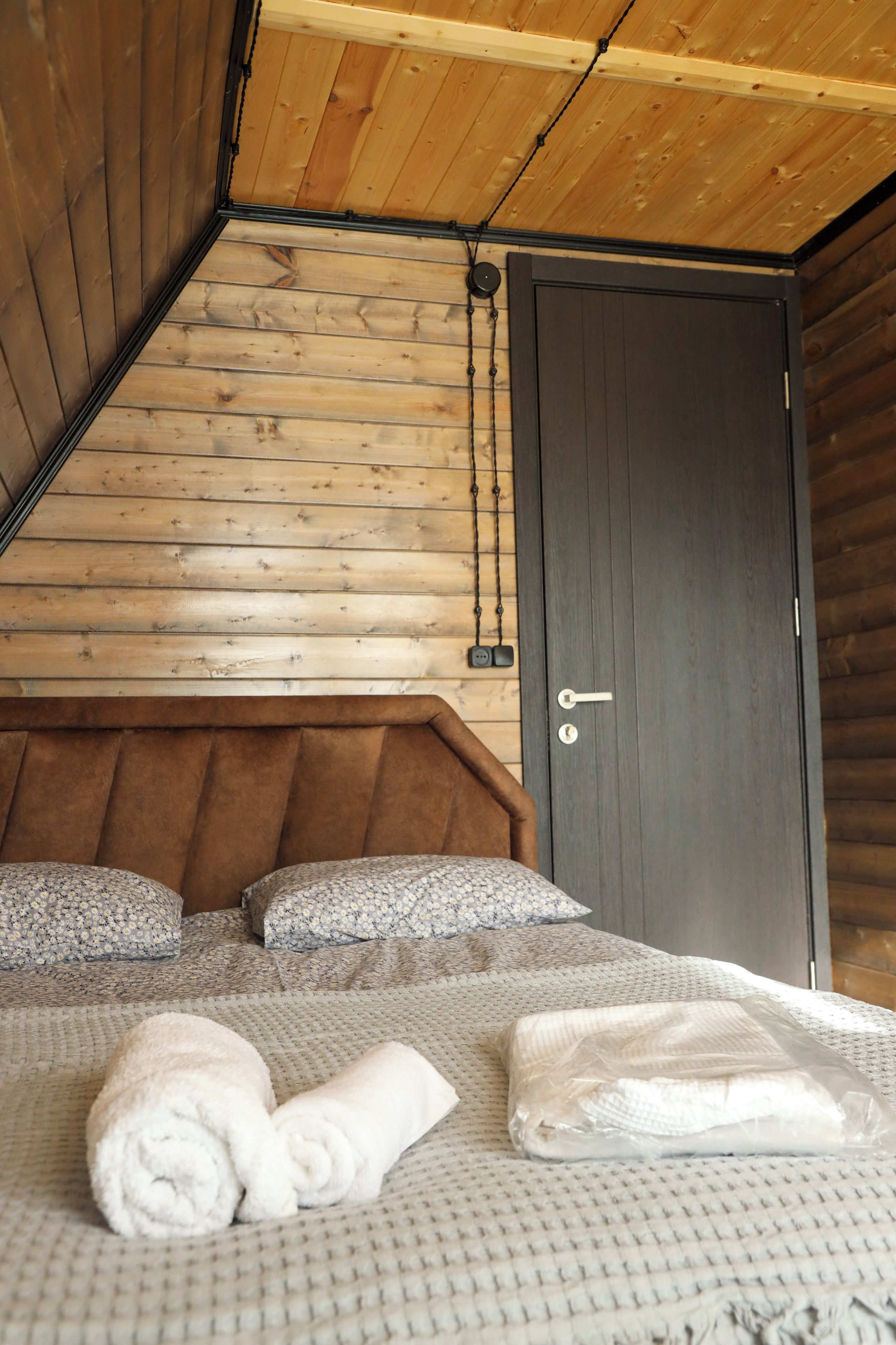 Ein Schlafzimmer einer Holzhütte mit schrägem Dach, einer dunklen Tür und einem Bett mit einem gemusterten Bettbezug und Kissen.