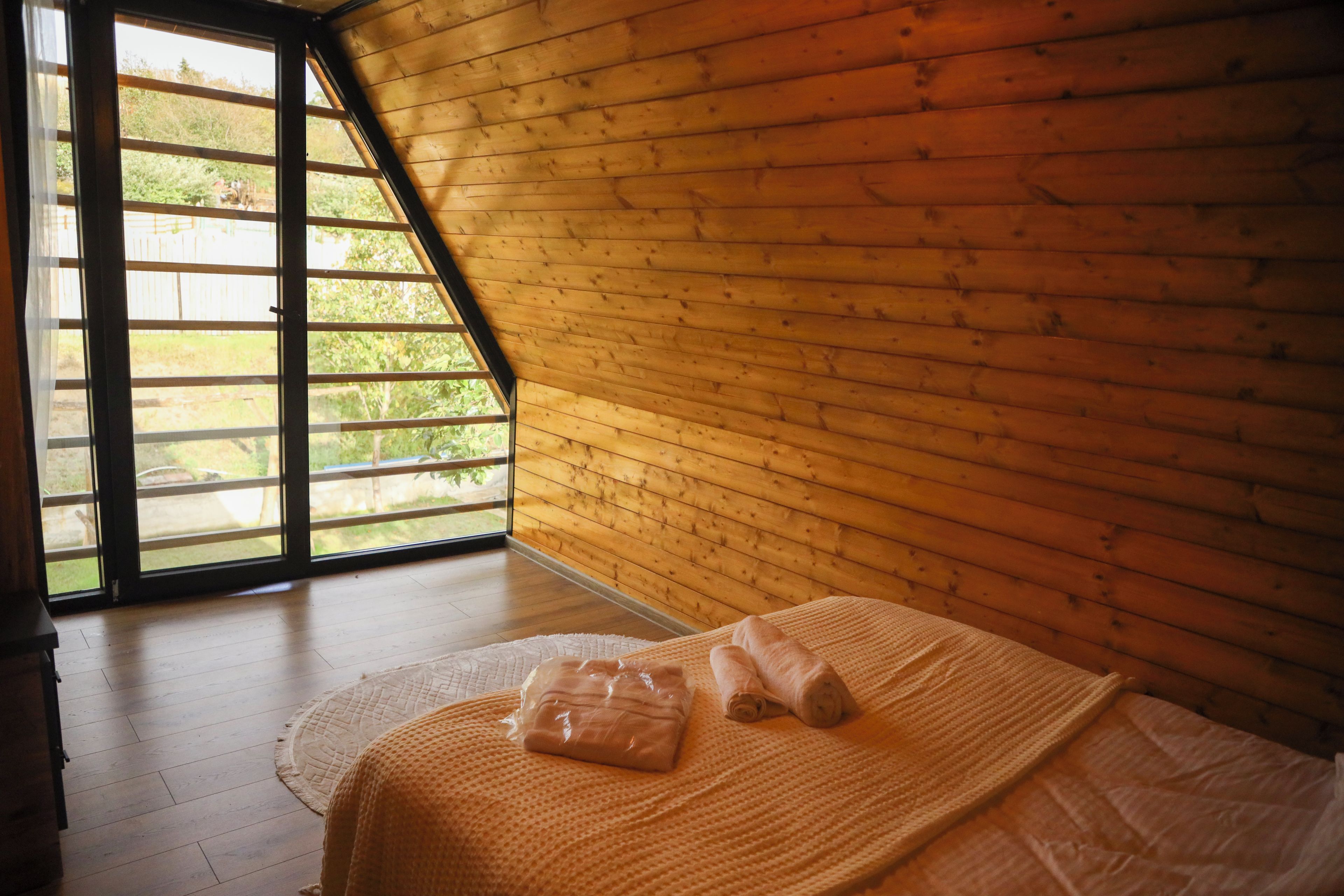 A room with a sloped ceiling, wooden walls, a bed with a white bedspread, and a window with a view of greenery.
