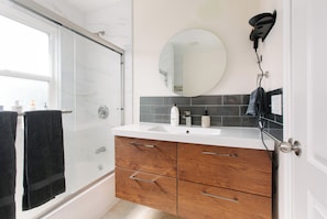 A bathroom with a glass shower enclosure, a white sink, wooden cabinets, a round mirror, and a hairdryer mounted on the wall. - California dreamin hilltop views! 4 bedrooms & 3 full baths (Los Angeles)