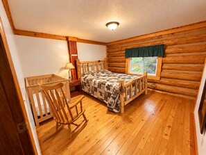 A log cabin bedroom with a wooden bed, a rocking chair, a nightstand, and a window with a view of trees. - Cranberry Mountain Lodge - Valemount (Valemount)