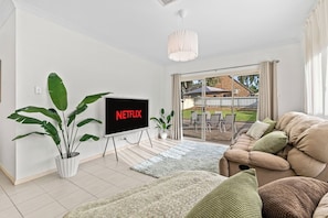 A living room with a flat-screen TV, a sofa, a potted plant, and a view through a glass door to an outdoor area. - Seednest | Spacious Magill BBQ Family Retreat (Magill)
