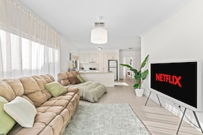 A modern living room with a sofa, a flat-screen TV, and a kitchen in the background. - Seednest | Spacious Magill BBQ Family Retreat (Magill)