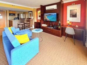 A living room with a sofa, a small round table, a TV mounted on the wall, and a kitchen area in the background. - @ Marbella Lane - Dreamy OceanView | Lanai Waikiki (Honolulu)
