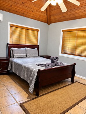 A bedroom with a wooden bed, two windows with blinds, a ceiling fan, and a tiled floor. - 3bed treat in West End with A/C, WiFi, parking. Chief's Haven (Roatan)