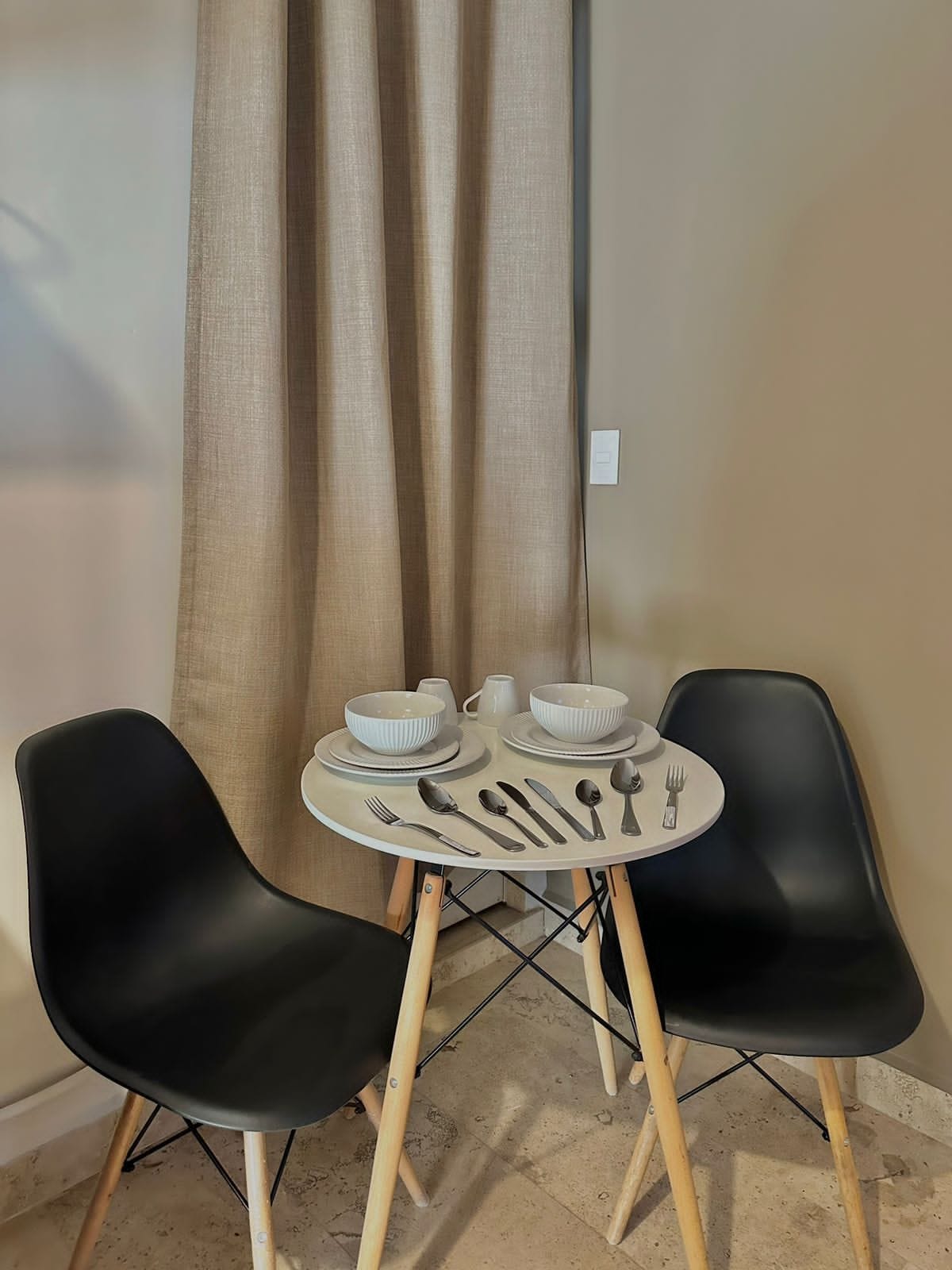 Uma pequena área de refeições com uma mesa redonda, duas cadeiras pretas e um conjunto de louça branca.