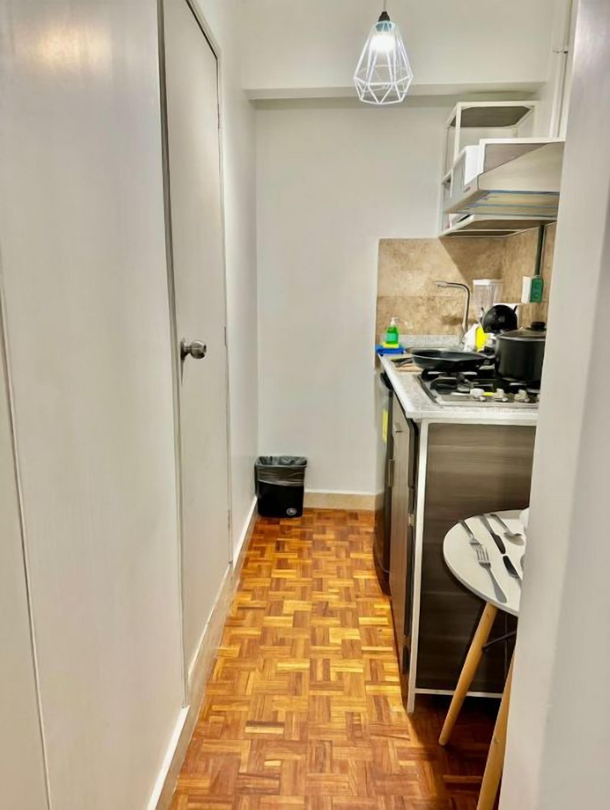 Un pasillo estrecho con suelo de parqué de madera que conduce a una cocina con estufa, lavamanos y una pequeña mesa de comedor.