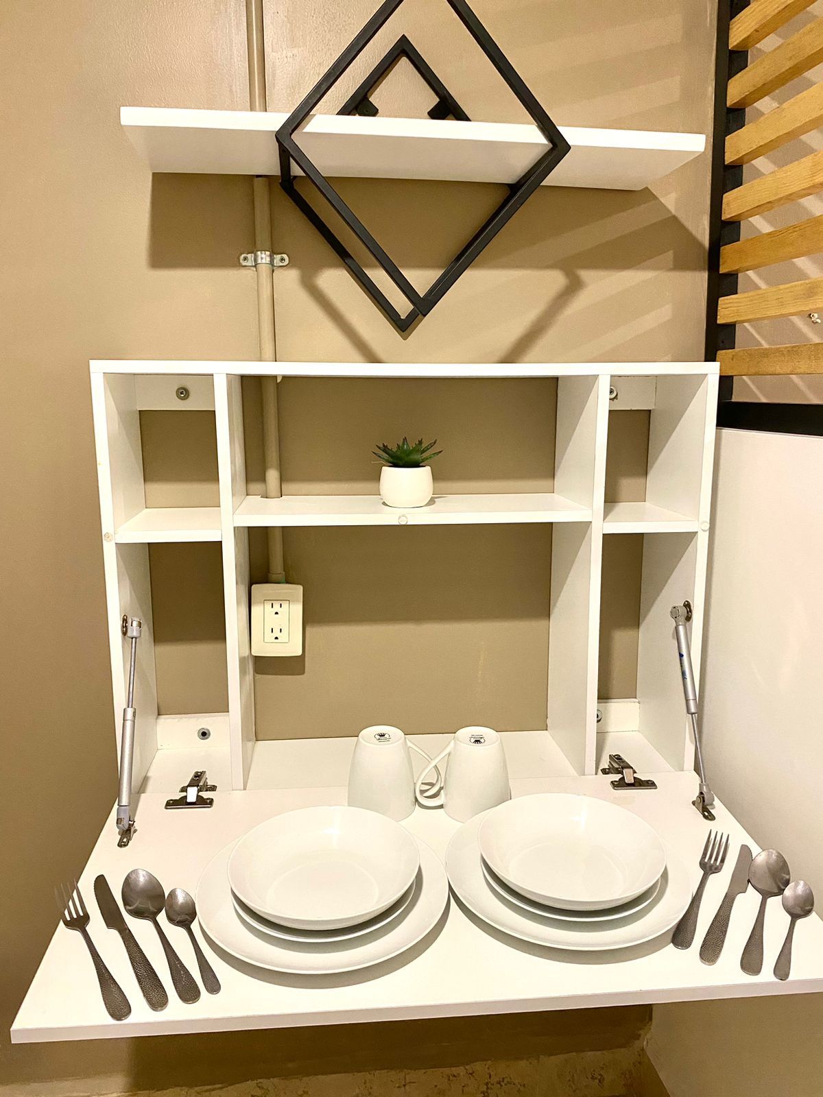 Un comedor con una mesa blanca dispuesta para dos personas, incluyendo platos, cubiertos y vasos. Hay una pequeña planta en maceta sobre la mesa.