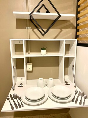 Un comedor con una mesa blanca dispuesta para dos personas, incluyendo platos, cubiertos y vasos. Hay una pequeña planta en maceta sobre la mesa.