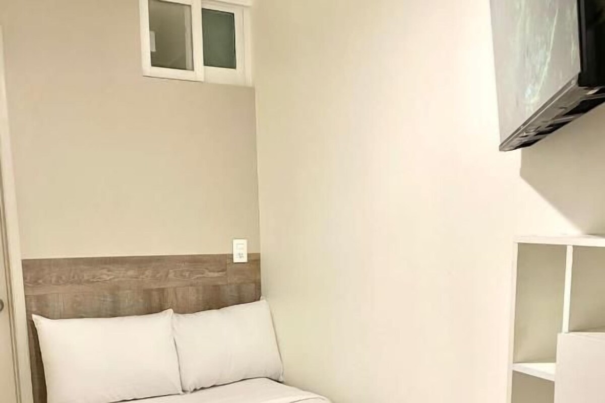 A single bed with a white comforter and a towel, a small window, and a television mounted on the wall.