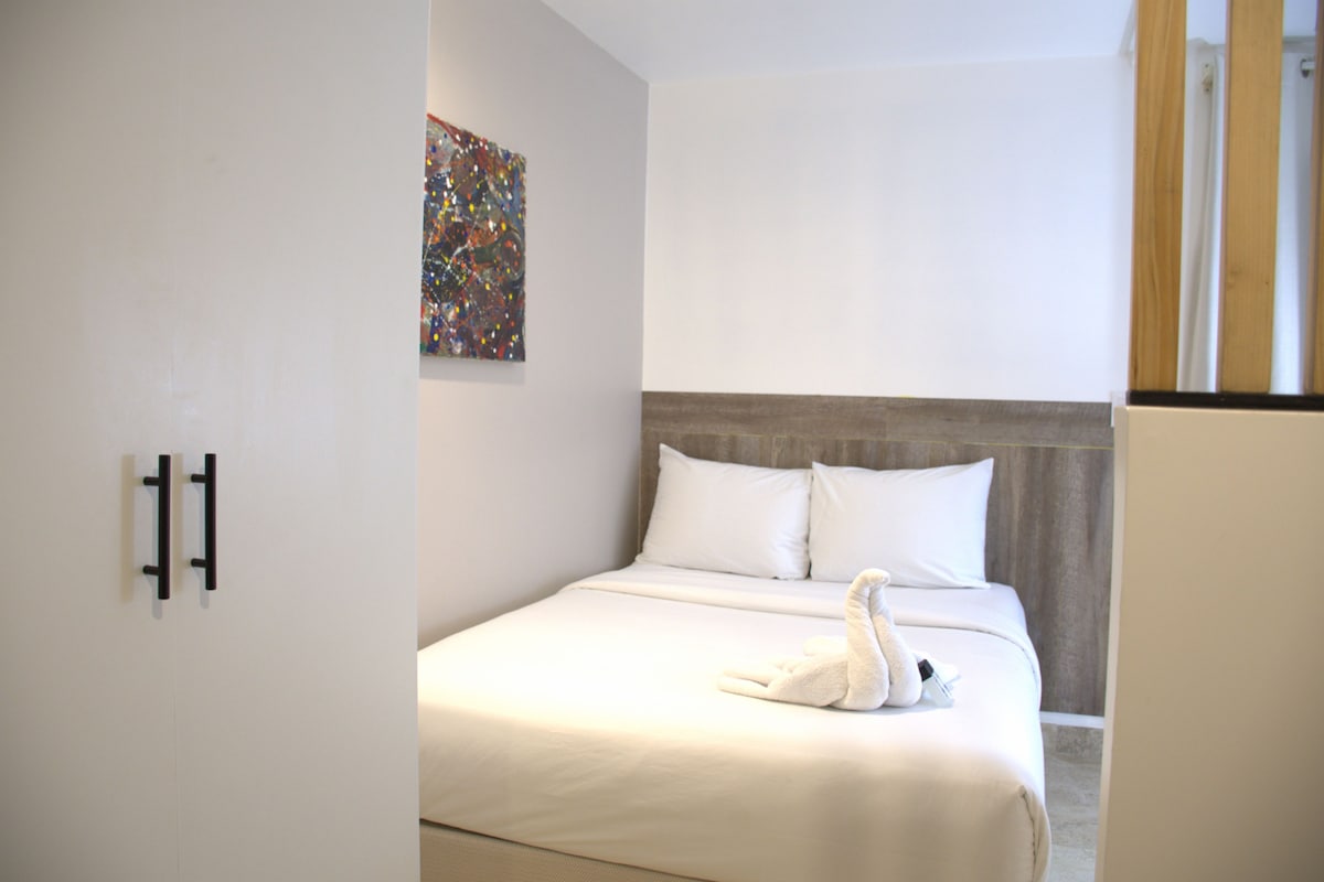 A hotel room with a bed, white pillows, a colorful abstract painting on the wall, and a towel on the bed.