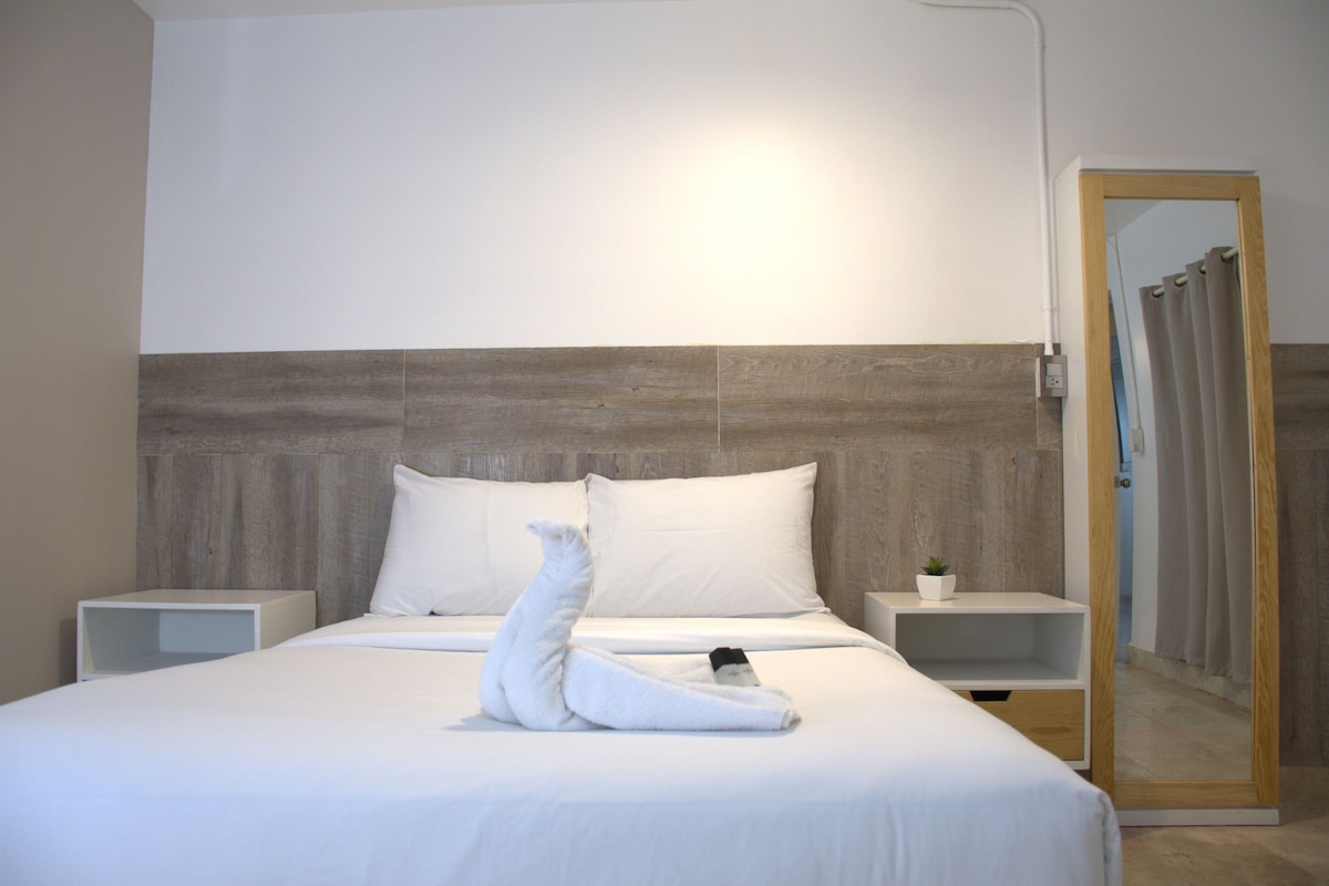 A hotel room with a large bed, two bedside tables, a wooden headboard, a mirror, and a door with curtains.