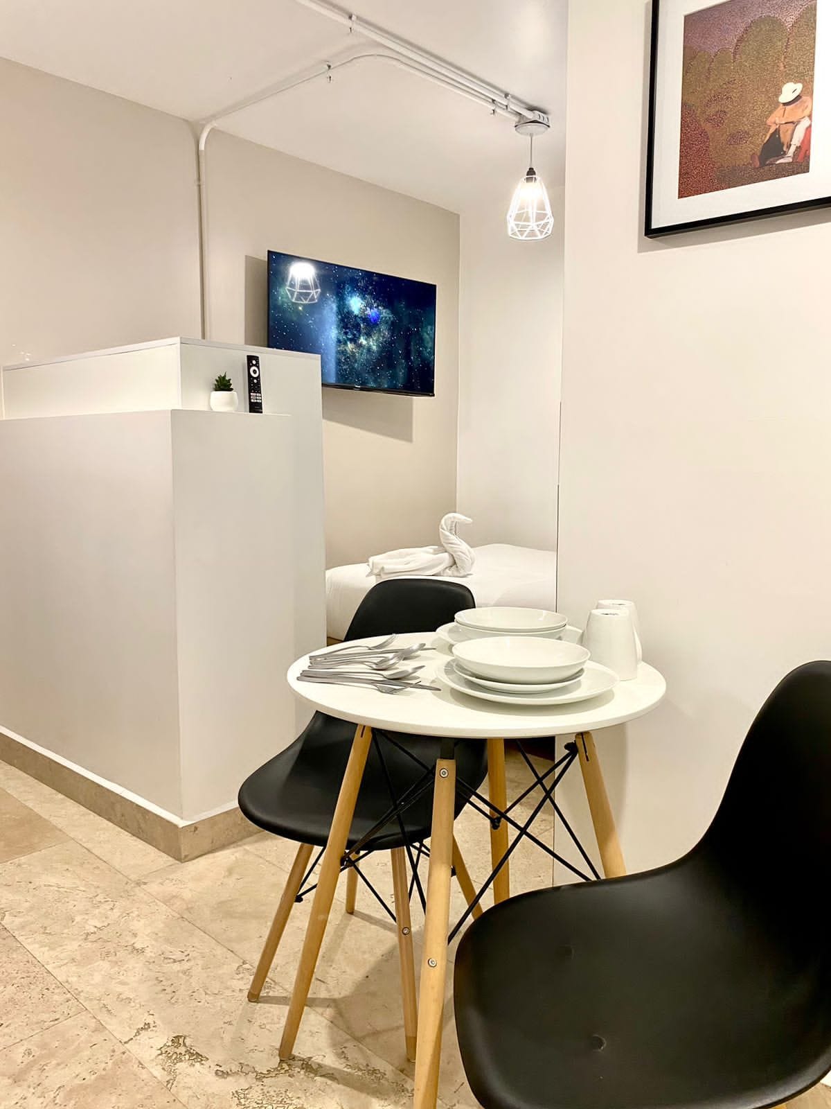 Une chambre d’hôtel compacte, avec une table à manger pour deux personnes, un téléviseur à écran plat fixé au mur et une œuvre d’art encadrée au-dessus du lit.