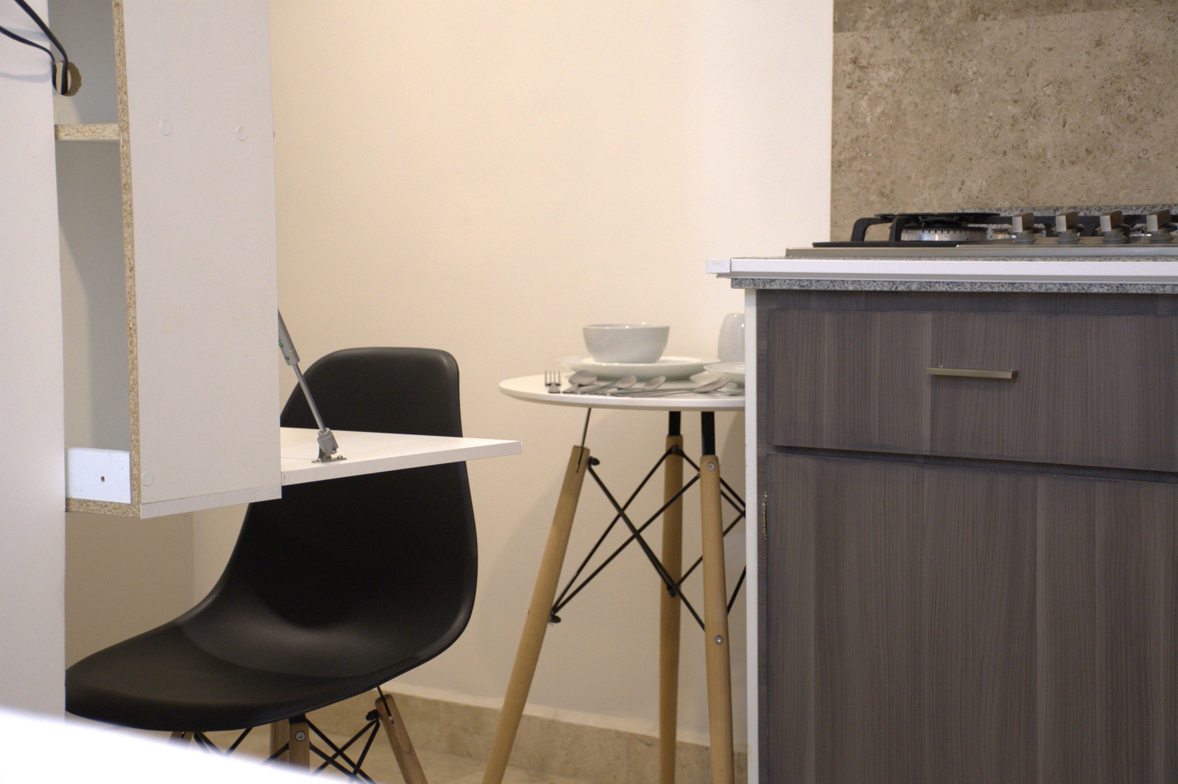 Cocina compacta con estufa a gas, una mesita redonda con un tazón y una silla negra.