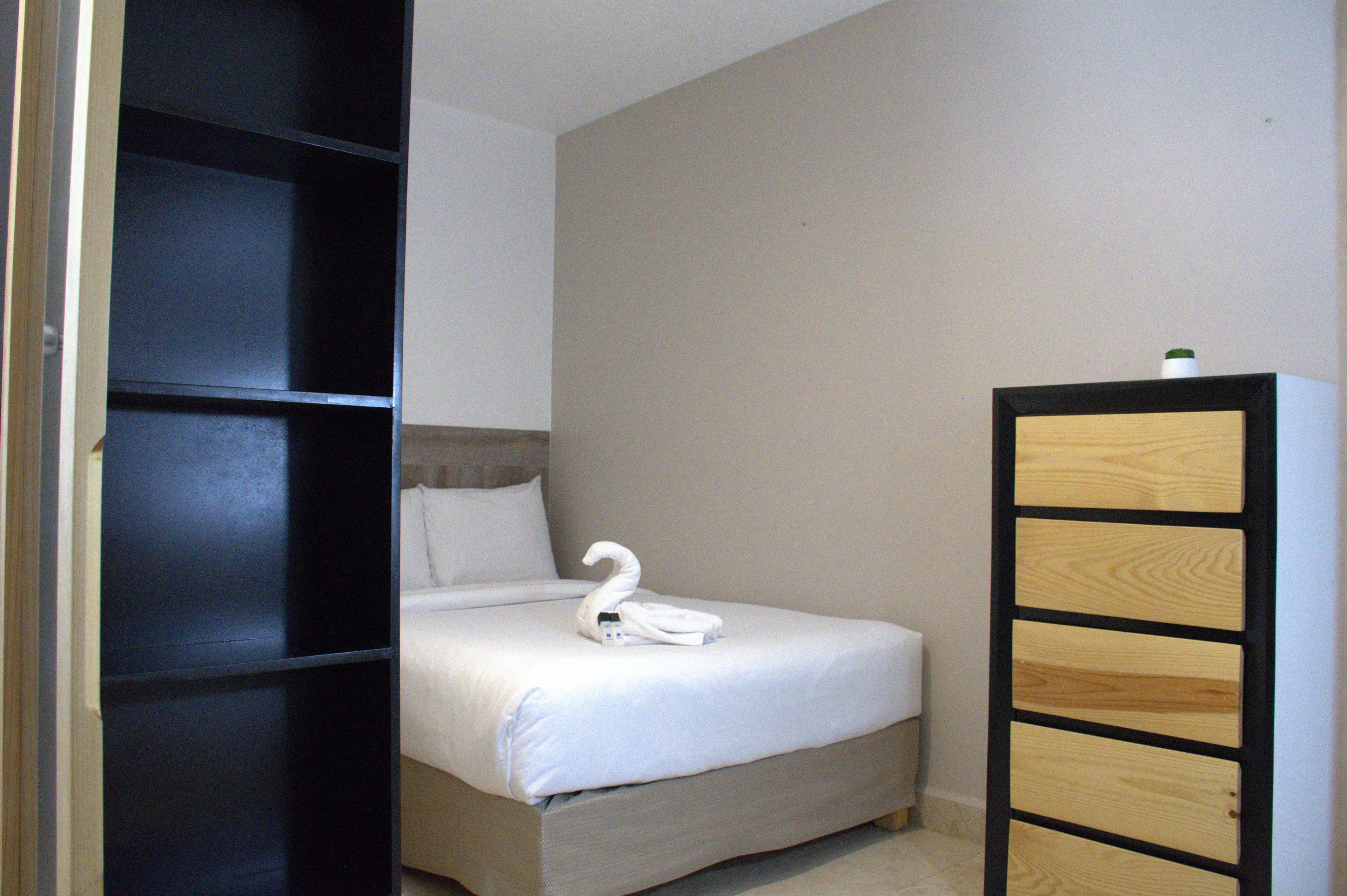 A single bed with a swan-shaped towel and a bottle of water, a wooden cabinet, and a partially open door revealing a dark room.