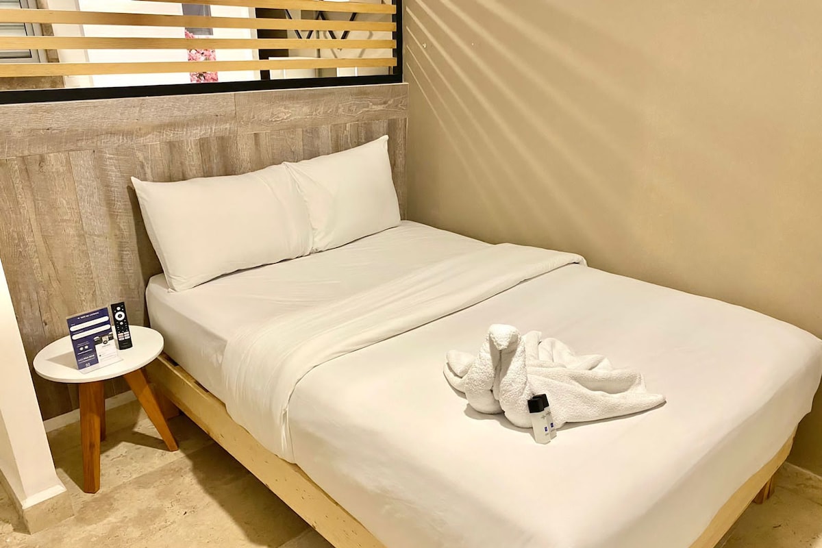 A single bed with white bedding, a wooden headboard, a small bedside table with a remote control, and a towel on the bed.