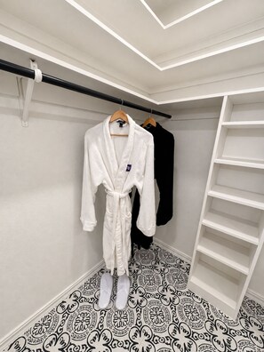 Un vestidor con un albornoz blanco colgado en una percha de madera, un albornoz negro y un estante blanco.