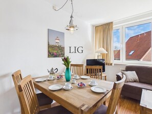 A dining area with a wooden table set for four, a TV, and a sofa in a room with a window. - Holiday Apartment Uhlenkamp 4 (Langeoog)