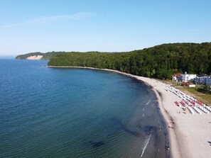 Ein Küstenstrand mit sandigem Ufer, eine Reihe von Strandsonnenliegen und ein Gebäude mit rotem Dach.