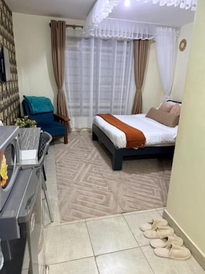 Ein kleines Hotelzimmer mit einem Bett, einem Schreibtisch mit Mikrowelle, einem Stuhl und einem Paar Hausschuhen auf dem Boden.