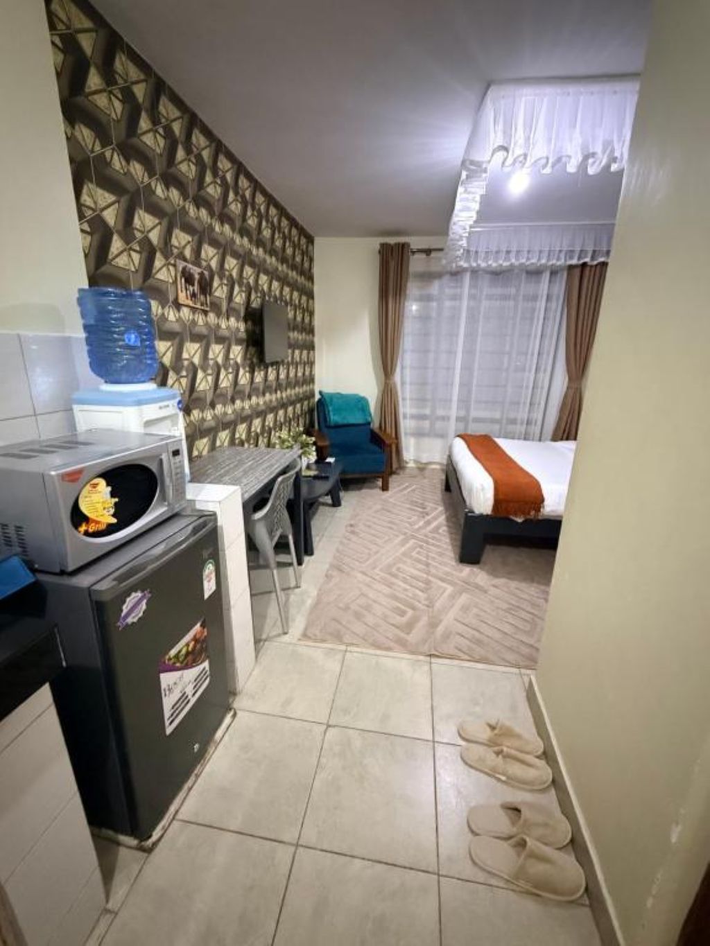 A compact hotel room with a kitchenette, a bed, a desk, and a large window with curtains.