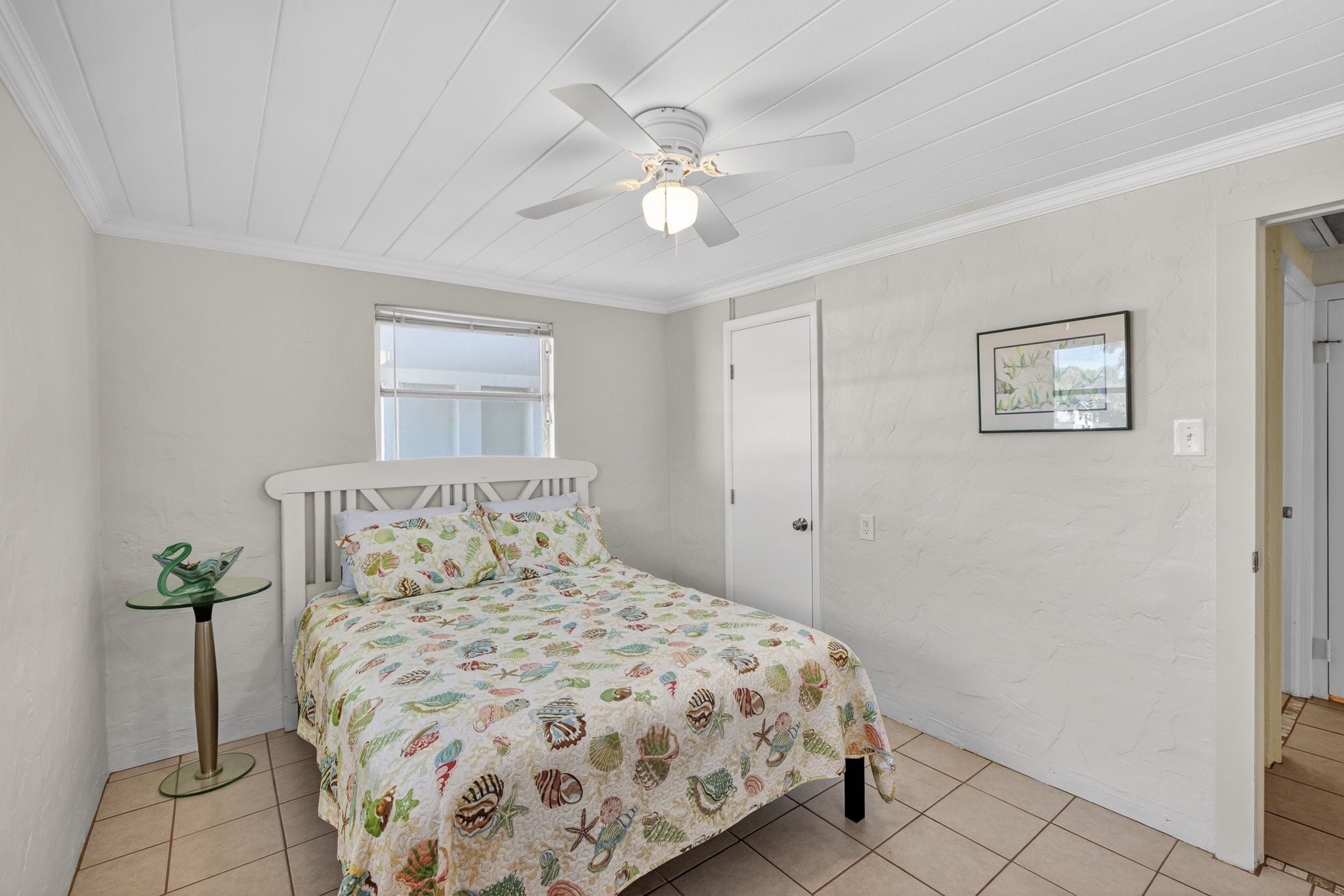 A bedroom with a bed, a ceiling fan, a window, a door, and a framed picture on the wall.