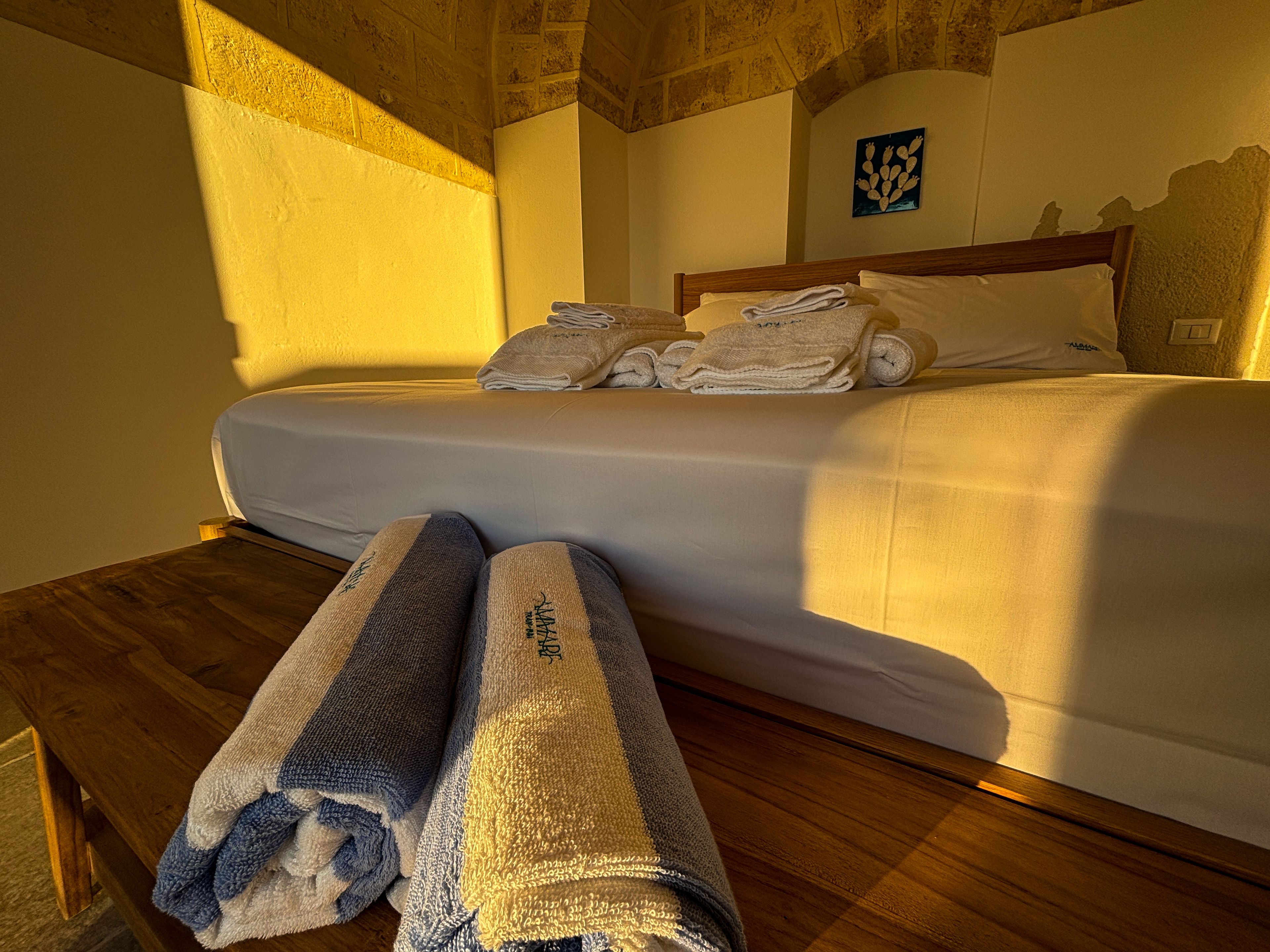 A hotel room with a bed, a wooden nightstand, and a large window allowing sunlight to enter.
