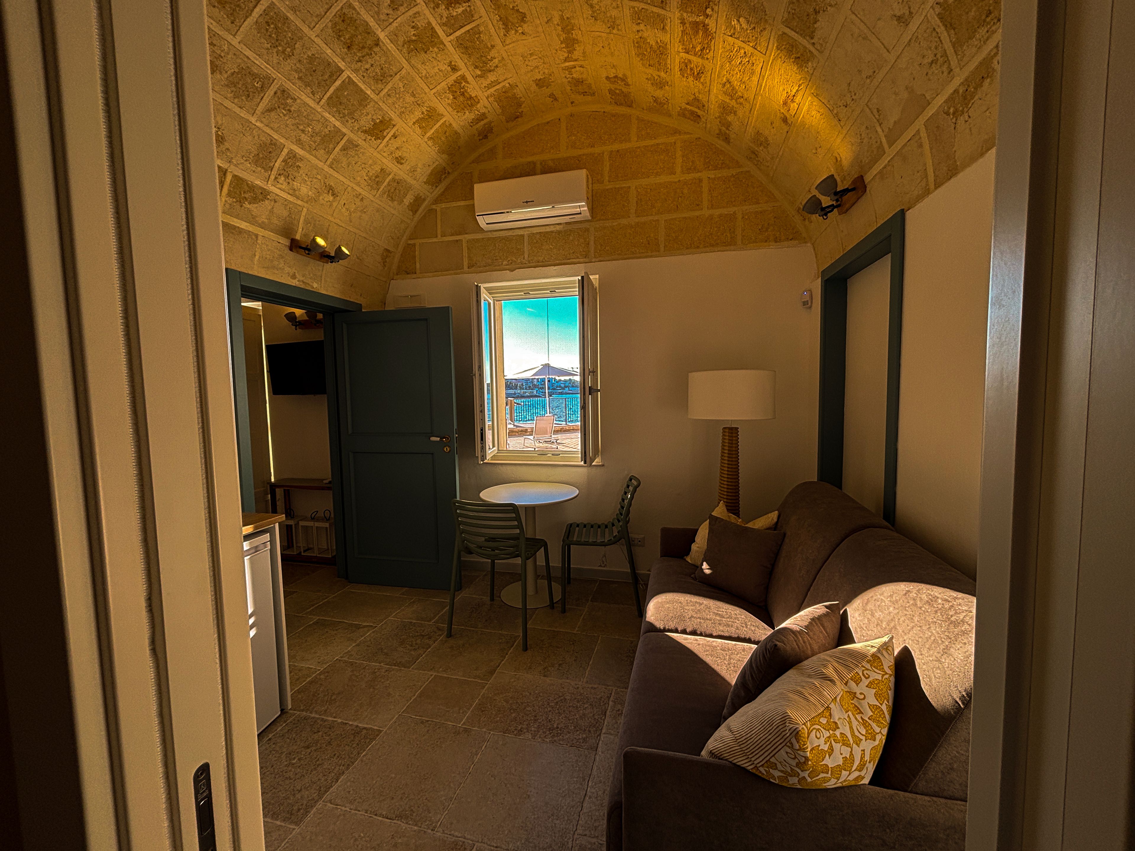 Una stanza accogliente con soffitto in pietra, una finestra vista, un divano con cuscini e un tavolo con sedie.
