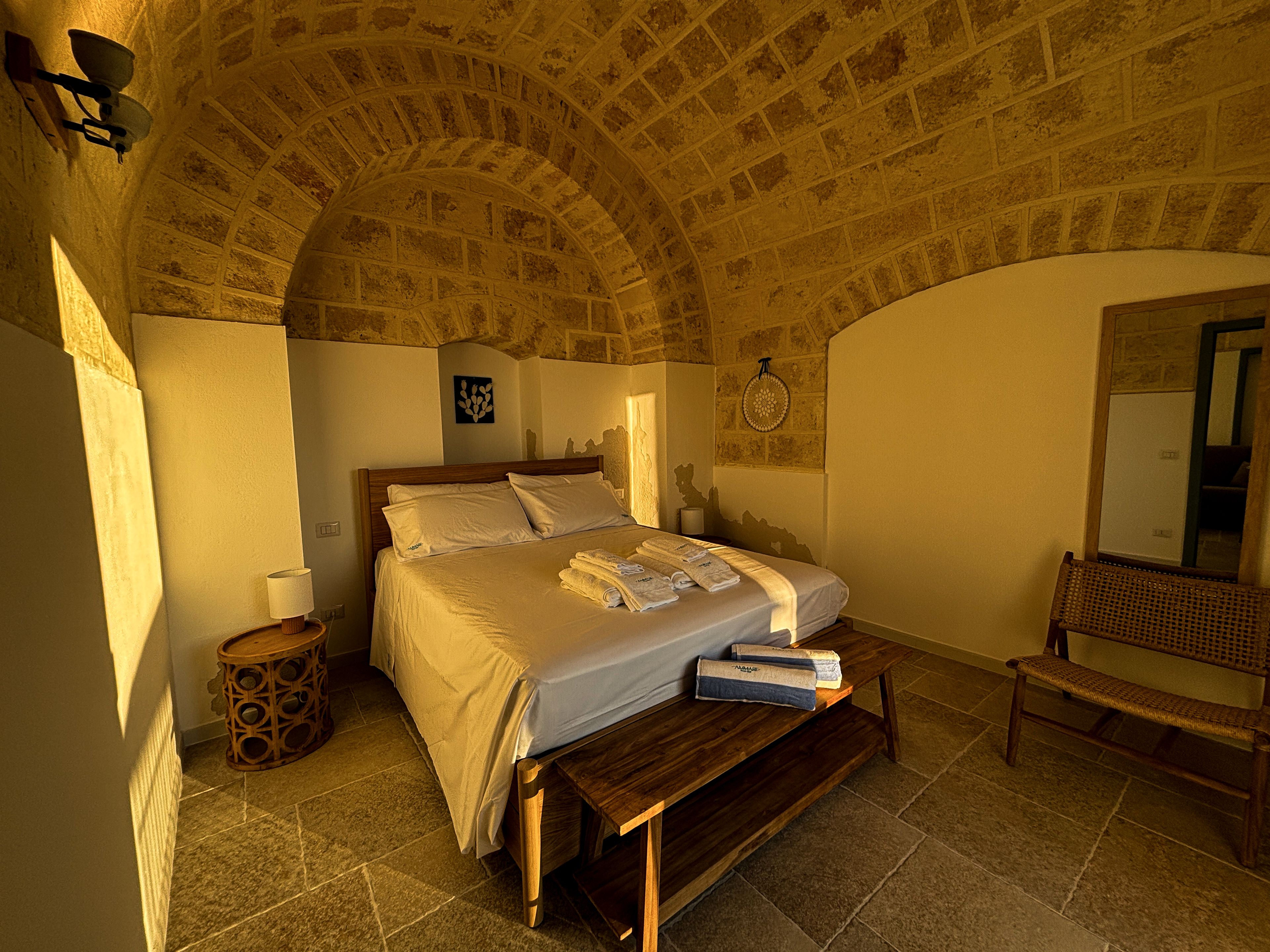 Una camera da letto con soffitto in pietra, un letto con lenzuola bianche, un comodino, una panca e uno specchio fissato al muro.