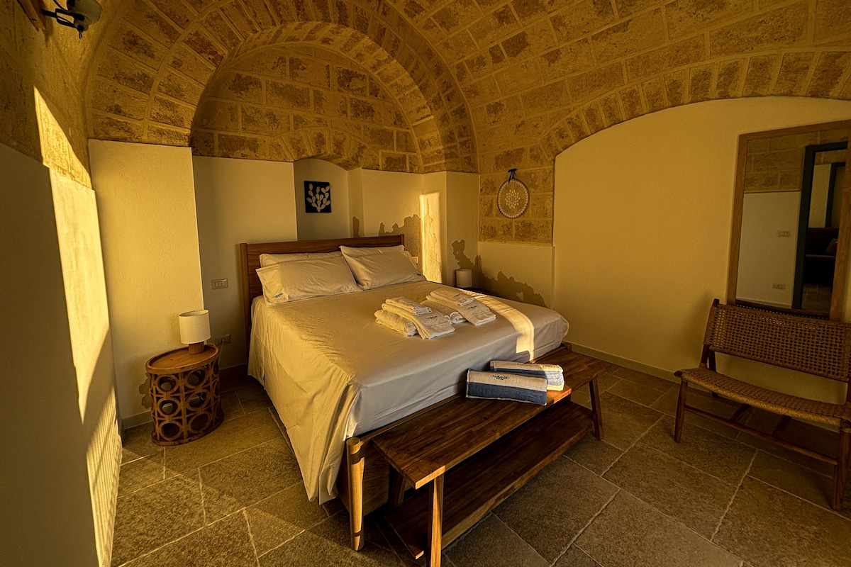 A bedroom with a stone ceiling, a bed with white linens, a bedside table, a bench, and a wall-mounted mirror.