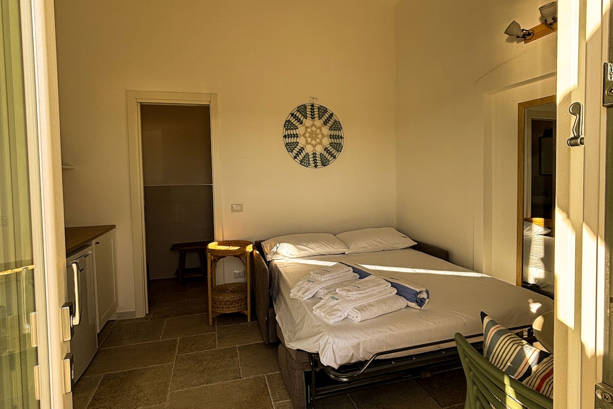 A bedroom with a bed, a bedside table, a wall decoration, and a view of the outside.