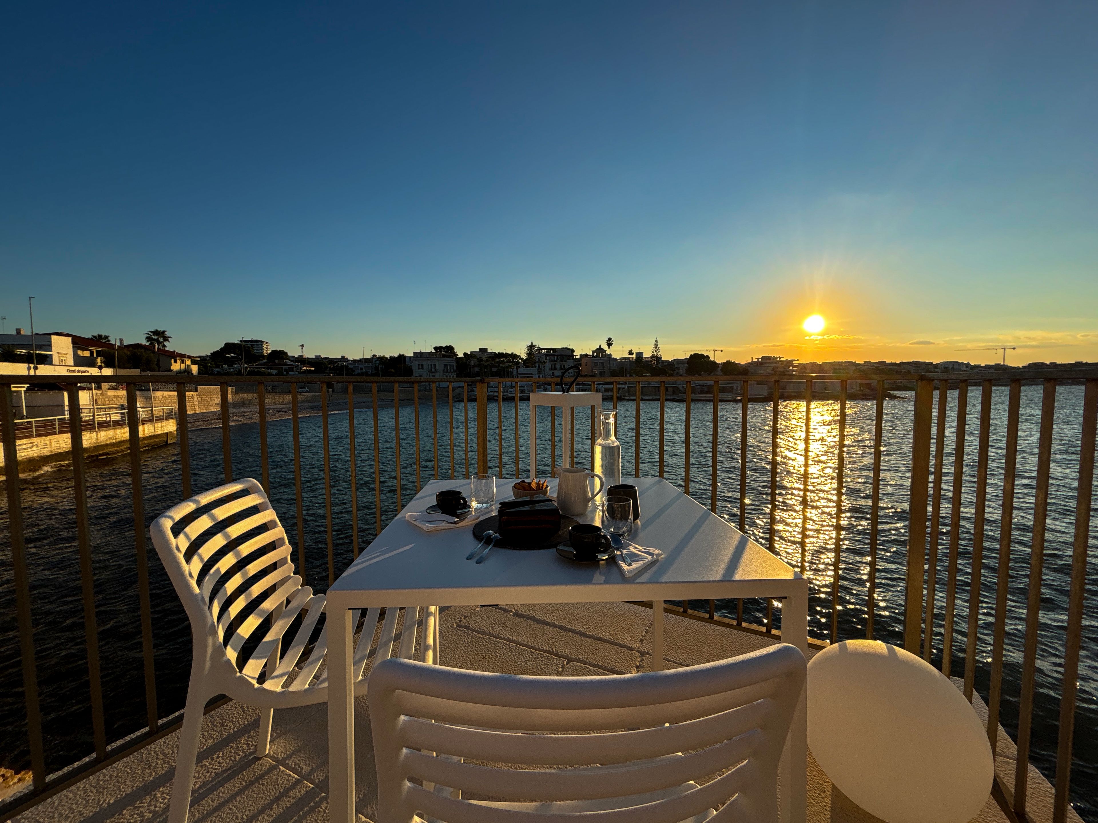 Een tafel met stoelen op een balkon met uitzicht op het water, met een zonsondergang op de achtergrond.