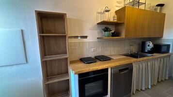 Cozinha compacta com forno embutido, micro-ondas e pia. Há uma prateleira de madeira acima do balcão e um pequeno vaso de planta na prateleira.