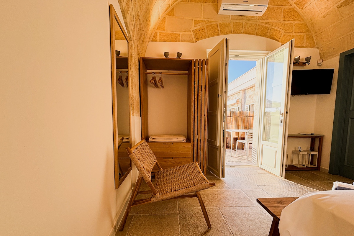 A hotel room with a stone wall, a wicker chair, a bed, and an open door leading to a balcony.