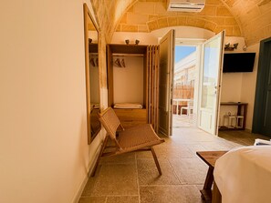 Une chambre d’hôtel avec un mur en pierre, un fauteuil en osier, un lit et une porte ouverte donnant sur un balcon.