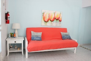 A room with a white sofa, two pillows with text, a painting, a small table with a lamp, and a fire extinguisher on the wall. - Casa Iliria 1: cozy 1-bedroom apartment with AC in charming Boquerón (Boquerón)