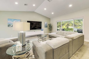 A modern living room with a sectional sofa, a glass coffee table, a flat-screen TV, and a large window with a view of greenery. - The Saltwater House at Deerfield (Deerfield Beach)