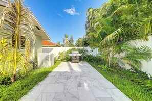 A paved pathway leading to a barbecue grill, surrounded by lush greenery and a white fence. - The Saltwater House at Deerfield (Deerfield Beach)
