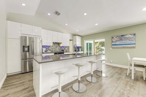 A modern kitchen with white cabinetry, a stainless steel refrigerator, and a bar area with stools. - The Saltwater House at Deerfield (Deerfield Beach)