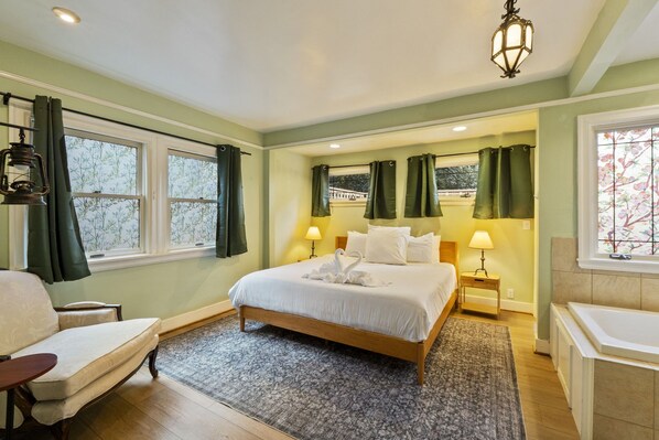 A hotel room with a bed, two bedside tables, a chair, a sofa, a bathtub, and a window with curtains. - Victorian 3BR with Jetted Tub + Fireplaces - Central District (Seattle)